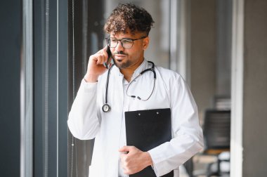 Beyaz önlüklü ve steteskoplu Arap erkek doktor elinde bir pano tutuyor ve hastane koridorunda bir telefon görüşmesi yapıyor.
