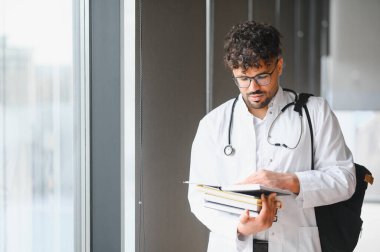 Beyaz önlüklü bir tıp öğrencisi, pencere kenarında ders çalışıyor, kitap ve notlar tutuyor, eğitimine ve gelecekteki kariyerine bağlılığını somutlaştırıyor.