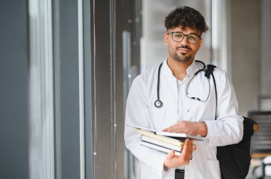 Beyaz önlük ve steteskop giyen genç Arap tıp öğrencisi, üniversite koridorunda kitap ve sırt çantası tutuyor.