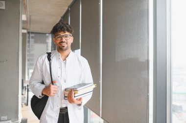Genç Arap tıp öğrencisi kitap taşıyor ve sırt çantası takıyor, ilham dolu parlak bir üniversite koridorunda güvenle yürüyor.