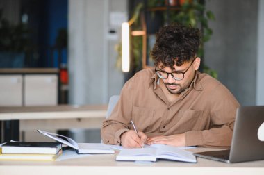 Genç Arap öğrenci, kitap ve teknoloji ile çevrili bir üniversite kütüphanesinde özenle çalışırken bir deftere notlar yazıyor.