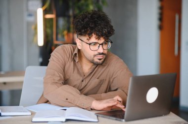 Bir üniversite kütüphanesinde kitaplarla çevrili dizüstü bilgisayarında yazarken konsantre olan genç Arap öğrenci, çevrimiçi öğrenimine odaklandı.