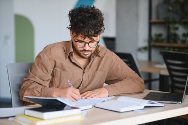 Genç Arap öğrenci üniversite kütüphanesinde okurken notlar yazıyor, sınavlara hazırlanıyor ve dizüstü bilgisayar kullanıyor.