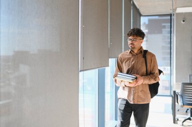 Bir üniversite koridorunda yürüyen Arap öğrenci, kitap ve sırt çantası taşıyor, öğrenme ve akademik takip ruhunu somutlaştırıyor.