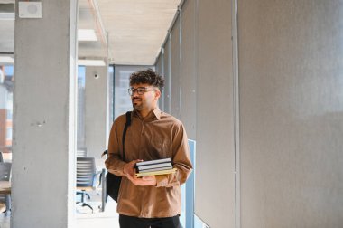 Genç Arap öğrenci üniversite koridorunda elinde kitaplar ve sırt çantasıyla yürüyor, gülümsüyor ve başka tarafa bakıyor.