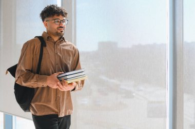 Genç Arap erkek üniversite öğrencisi kolej koridorunda ders aralarında pencere ışığında yürüyen kitaplar taşıyor.