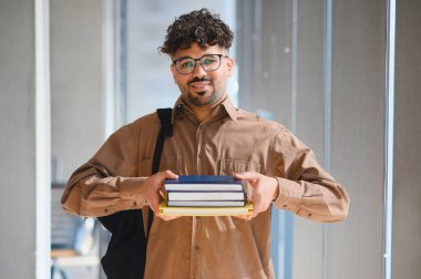 Genç Arap öğrenci kitap taşıyor ve parlak bir üniversite koridorunda güvenle yürüyor, akademik başarının ruhunu somutlaştırıyor.