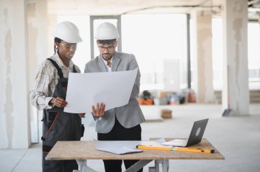 Mimar ve müteahhit başlık takıyor ve bir inşaat alanının planlarını inceliyor, bina planlarını tartışıyor ve proje ilerlemesini tartışıyorlar.