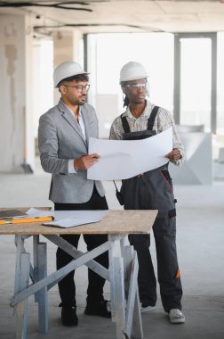 Mimar ve müteahhit inşaat halindeki bir binanın masasında dururken planlarını gözden geçiriyorlar.