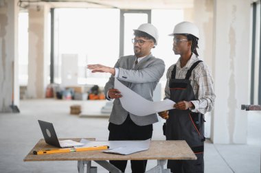 Güvenlik kaskı takan mimar ve müteahhit, hareketli bir inşaat alanının planları hakkında bir tartışmaya girdi, proje planları üzerinde işbirliği yaptı.