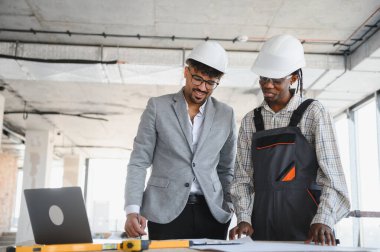 Güvenlik kaskı takan mimar ve müteahhit, bir inşaat alanının planlarını inceliyor, işleri için bir dizüstü bilgisayar ve ruhsal seviye kullanıyor.