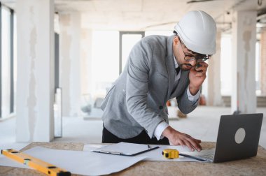 Arap mimar takım elbise giyiyor ve inşaat projesini yönetiyor, dizüstü bilgisayar ve akıllı telefon kullanıyor, planlar ve masa üzerindeki ölçüm araçlarıyla