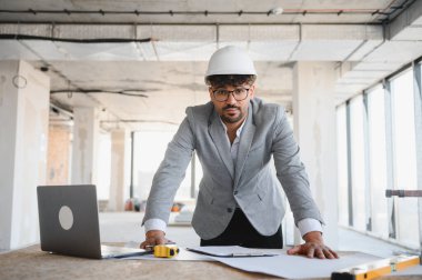 Arap mimar, inşaat projesinin içerisindeki planlar ve dizüstü bilgisayarla masaya yaslanarak inşaat projesini denetliyor.