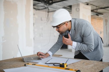 Arap mimar inşaat sahasında çalışırken telsiz ve dizüstü bilgisayar kullanarak planlara bakar ve hesaplamalar yapar.