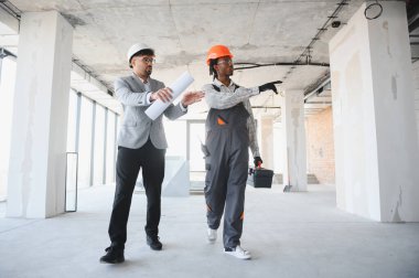 İnşaat alanından geçen inşaatçıyla inşaat sürecini tartışan mimar elinde planlar tutuyor