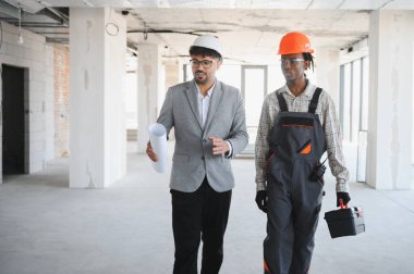 Mimarın elinde inşaat aşamasını tartışan planlar var. İnşaat aşamasında bina içinde yürüyen bir alet çantası taşıyan inşaatçıyla.