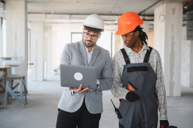 Mimarın elinde bina teftişi sırasında baret ve tulum giyen inşaatçının planlarını gösteren dizüstü bilgisayar var.