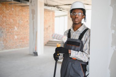 Genç Afro-Amerikan inşaat işçisi elinde boya silindiri, güvenlik gözlüğü ve şapka takıyor inşaat halindeki bir binada duruyor.