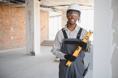 Üniformalı ve koruyucu şapkalı genç Afrikalı Amerikalı müteahhit inşaat alanında ruh seviyesini koruyor.