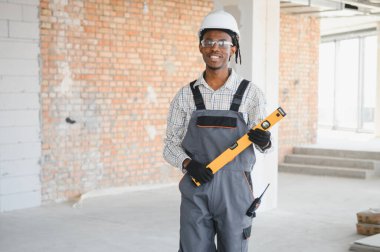Afrikalı Amerikalı inşaat işçisi bir inşaat sahasında koruyucu gözlük ve şapka takarak ruh seviyesinde duruyor.