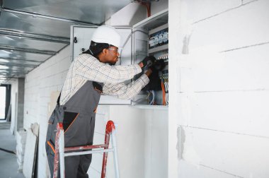 Merdivendeki elektrikçi inşaat halindeki bir binanın elektrik paneli üzerinde çalışıyor. Elektrik tesisatı ve elektrik güvenliği sağlanıyor.