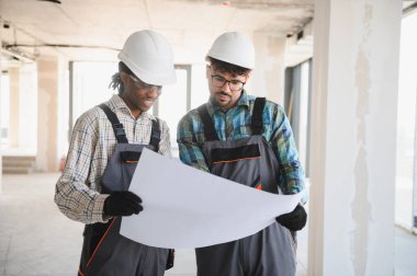 İki inşaat işçisi inşaat halindeki bir binanın mimari planlarını gözden geçiriyorlar, takım çalışmalarını ve çeşitliliğini vurguluyorlar.