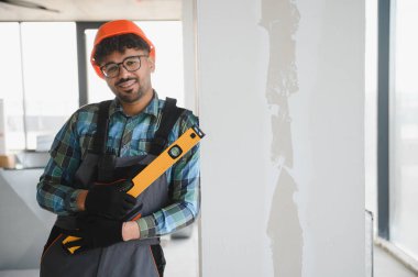 Gülümseyen Arap mühendis inşaat alanında bir duvarın yanında dururken kasketli ve ruh seviyesini koruyor.