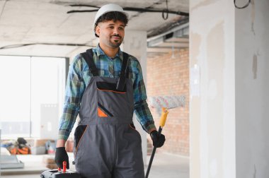 Gülümseyen Arap mühendis boya silindiri ve alet çantası taşıyor, yoğun bir inşaat sahasında güvenle yürüyor, yenileme konusunda uzmanlık gösteriyor