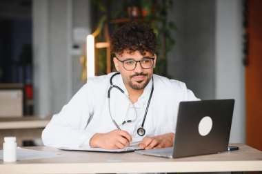 Laboratuvar önlüğü ve steteskop giyen odaklanmış erkek doktor laptopunu kullanıyor ve tıp bürosunda online danışmanlık sırasında notlar alıyor.