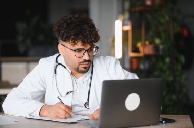 Laboratuvar önlüğü ve steteskop giyen odaklanmış erkek doktor, tıp bürosunda online danışmanlık sırasında dizüstü bilgisayar kullanıyor ve notlar yazıyor.