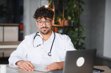 Laboratuvar önlüğü ve steteskop giyen kendine güvenen Arap erkek doktor modern tıp ofisinde dizüstü bilgisayar kullanarak masada oturuyor.