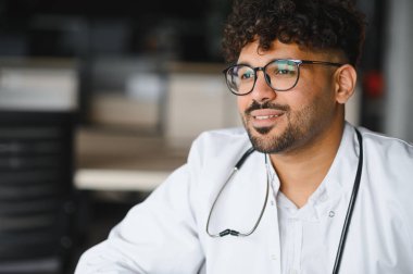 Gözlük takan, steteskoplu laboratuvar önlüğü giyen, gülümseyen ve hastane ortamında yüzünü çeviren kendine güvenen Arap erkek doktor.