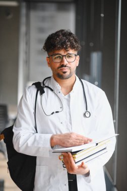 Genç Arap tıp öğrencisi elinde kitaplarla beyaz önlük ve steteskop giyiyor ve gözlerini kaçırıyor.