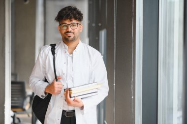 Kitap ve sırt çantası taşıyan kendine güvenen tıp öğrencisi modern bir üniversite koridorunda adım adım yürüyor, azim ve hırsı somutlaştırıyor.