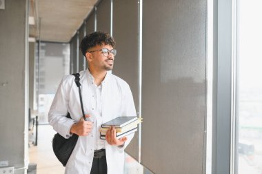 Genç tıp öğrencisi üniversite koridorunda elinde kitap ve sırt çantasıyla pencereden dışarı bakıyor.