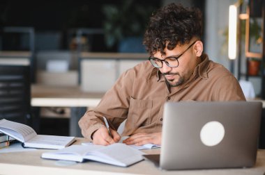Genç Arap öğrenci laptoptan notlar alıyor, parlak bir üniversite kütüphanesinde çalışmalarına odaklanmış.