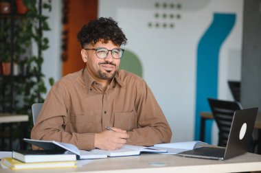 Üniversite kütüphanesinde dizüstü bilgisayarla çalışırken not alan odaklanmış Arap öğrenci, sınavlara hazırlanıyor ve araştırma projesi üzerinde çalışıyor