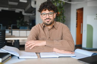 Açık kitaplarla bir masada oturmuş parlak bir üniversite kütüphanesinde sınavlara hazırlanan odaklanmış bir öğrencinin portresi.