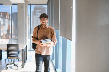 Genç Arap öğrenci bir üniversite koridorunda yürüyor, elinde kitaplarla gülümsüyor, güven ve hırs saçıyor.