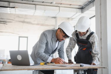 Mimar ve inşaatçı baret takıyor. Gün ışığında hareketli bir inşaat sahasının ayrıntılı planlarını birlikte gözden geçiriyorlar.