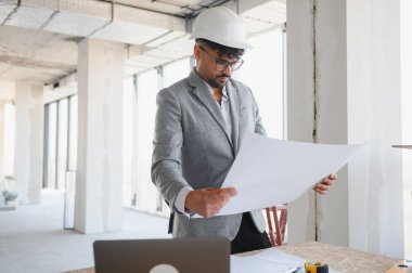 Arap mimar inşaat alanının planlarını inceliyor, başlık ve gözlük takıyor, proje detayları ve tasarıma odaklanıyor.