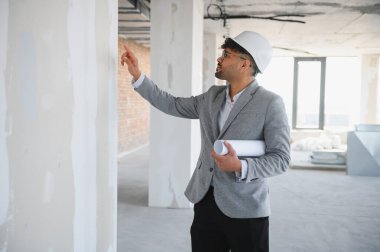 Arap mimar, inşaat alanındaki gelişmeleri incelerken, elinde planlar tutarken duvara bakan kalın şapka ve takım elbise giyiyor.