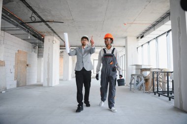 Mimarın elinde planlar var ve inşaat halindeki bir binanın içindeki inşaatçıyla inşaat sürecini tartışıyor.