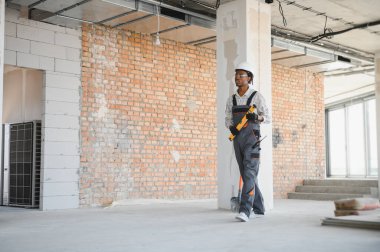 Afrika kökenli Amerikalı inşaat işçisi inşaat halinde, ruh seviyesini koruyor, iş tulumu, şapka ve eldiven giyiyor.