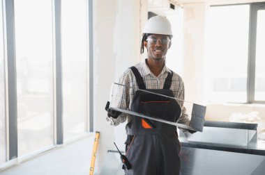 Gülümseyen Afro-Amerikan inşaat işçisi inşaat alanında güvenlik kaskı ve eldiven takarak bir havalandırma sistemi parçası taşıyor.