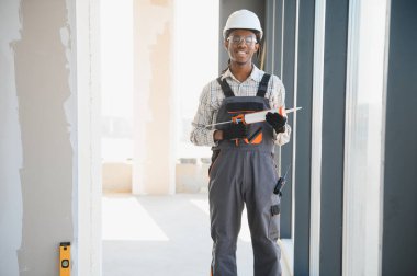 Gülümseyen Afro-Amerikan inşaat işçisi inşaat halindeki binanın penceresinin yanında dikilirken silikon dolgu tabancası tutuyor.