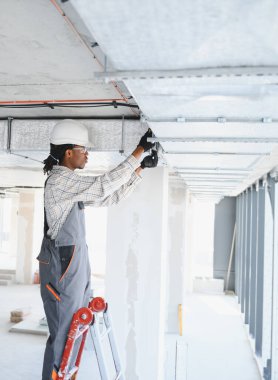 Afrikalı Amerikalı inşaat işçisi merdivene havalandırma boruları yerleştiriyor, inşaat halindeki bir binada tavan havalandırma sistemi üzerinde çalışıyor.