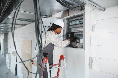 Merdivendeki elektrikçi inşaat halindeki bir binaya elektrik paneli yerleştiriyor, düzgün elektrik tesisatı ve güvenlik önlemleri sağlıyor