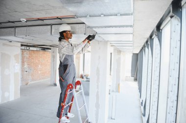 Genç Afro-Amerikan inşaat işçisi inşaat halindeki bir binanın merdiveninde dururken tavana havalandırma sistemi kuruyor.