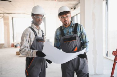 İki inşaatçı, baret ve tulum giyiyorlar, hareketli bir inşaat alanının ayrıntılı planlarını yakından inceliyorlar, proje planlarını tartışıyorlar.